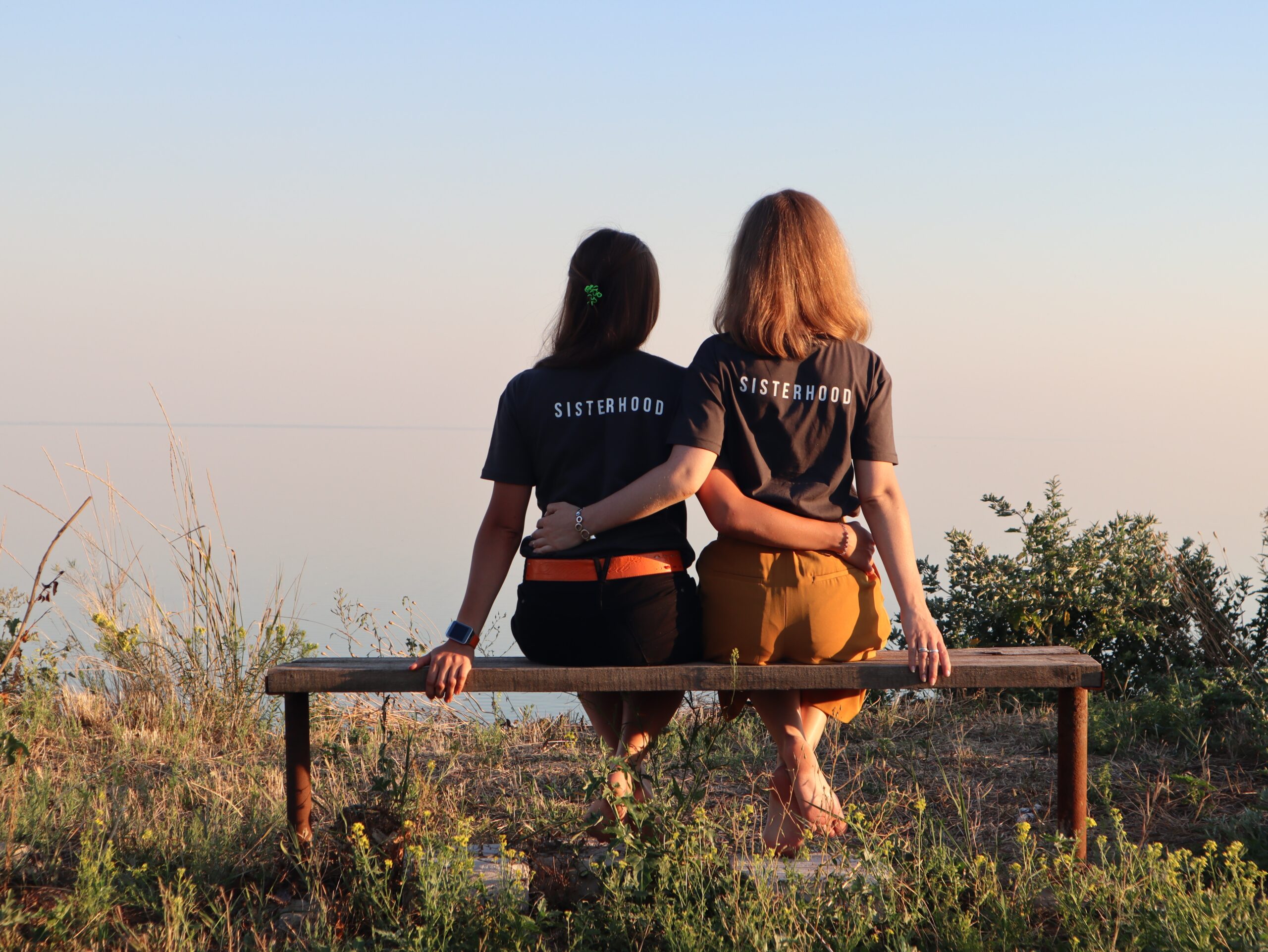 2 girls sitting on a bench in the park with their backs to us