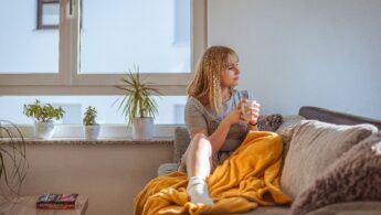 Girl sitting cozily on a sofa with a cup of tea stirring out the window.