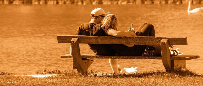 man and woman with a phone in hand siting on a bench in a park 